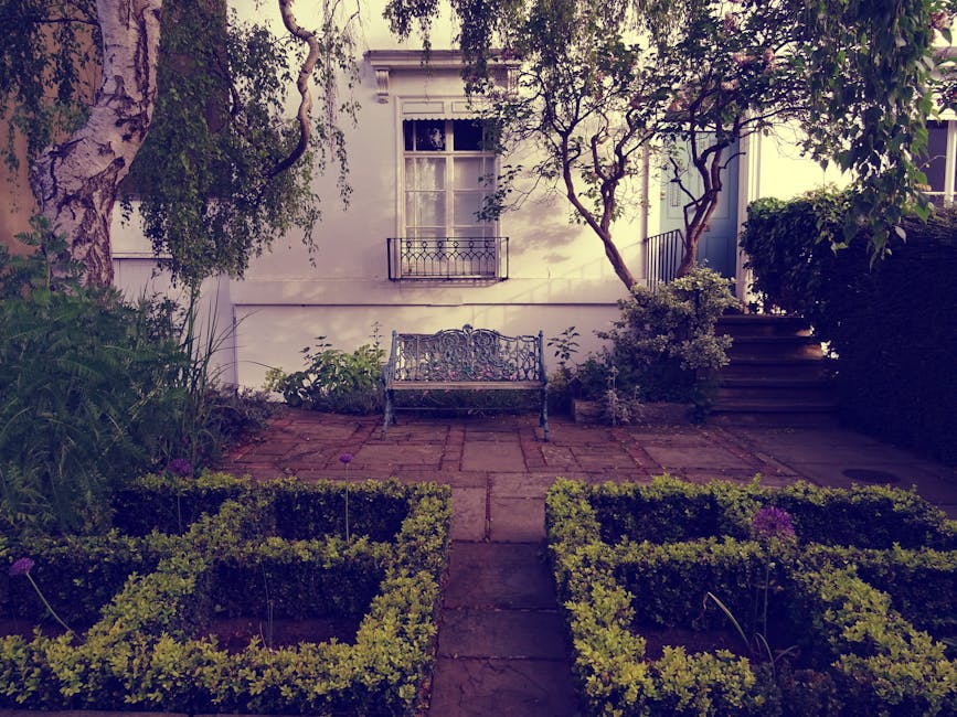 A small, neatly maintained garden courtyard featuring a paved brick patio with a decorative wrought iron bench positioned centrally against a white exterior wall of a residential building. The garden